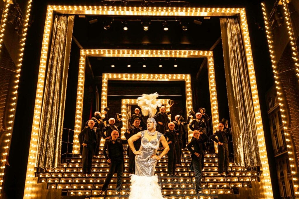 Zaza en robe à paillettes et coiffe de plumes blanches au centre d'un escalier illuminé, entourée de la troupe, dans La Cage aux Folles
