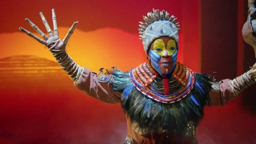 Rafiki, bras levés et visage peint, devant un ciel de savane orangé dans Le Roi Lion au Théâtre Mogador