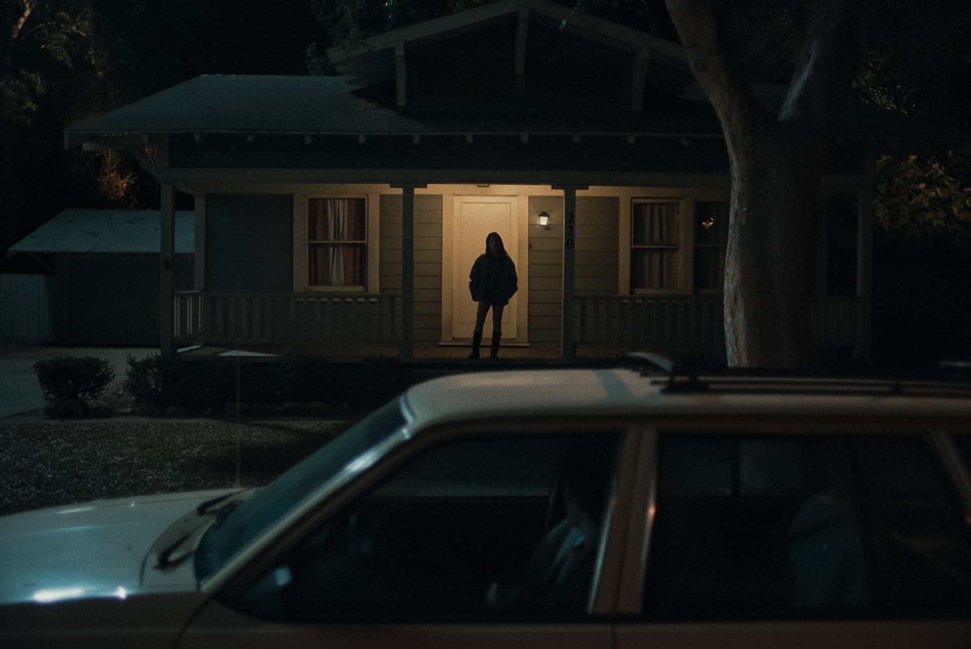Scène nocturne d'Obsession de Curry Barker : une silhouette immobile se découpe dans l'encadrement d'une porte éclairée, une voiture garée au premier plan