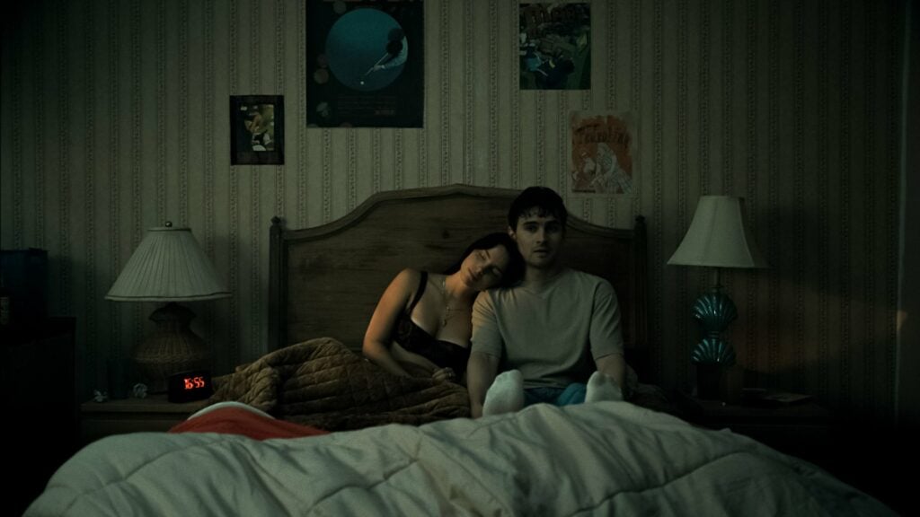 Inde Navarrette et Michael Johnston dans Obsession de Curry Barker : Nikki, blottie contre Bear, assis dans un lit dans une chambre à la lumière tamisée